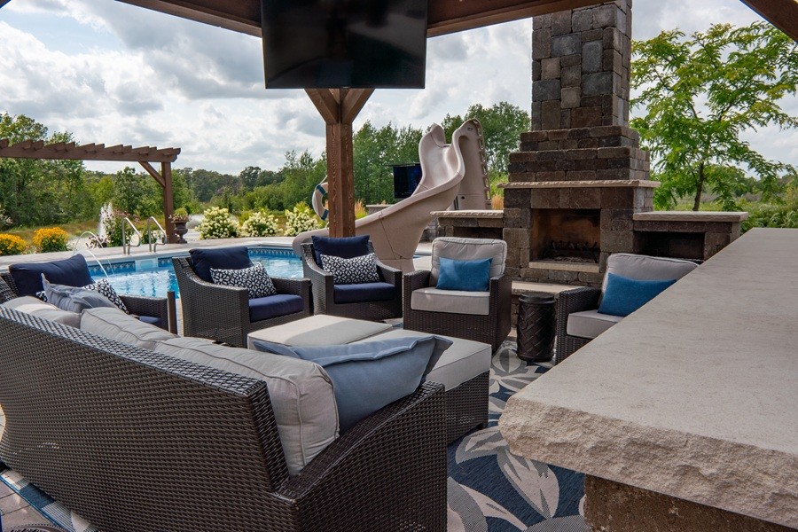 Outdoor living space with wicker seating, a fireplace, mounted TV, and pool with a slide, surrounded by lush landscaping and trees.